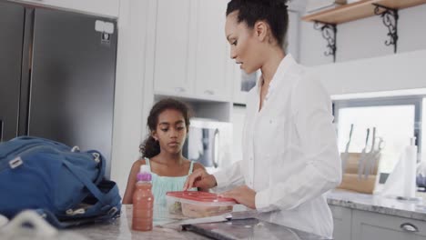 Happy-african-american-mother-and-daughter-packing-lunch-for-school-in-kitchen,-slow-motion
