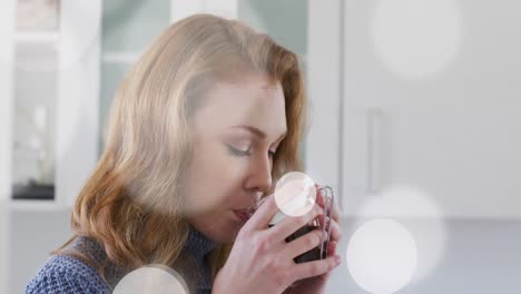 Animation-of-light-spots-over-caucasian-woman-drinking-coffe-in-kitchen