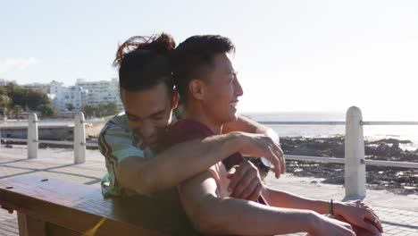 Happy-diverse-gay-male-couple-sitting-on-bench-and-embracing-at-promenade-by-the-sea,-slow-motion