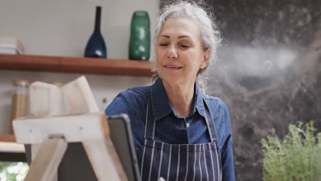Happy-senior-caucasian-woman-using-tablet-and-cooking-in-kitchen,-slow-motion