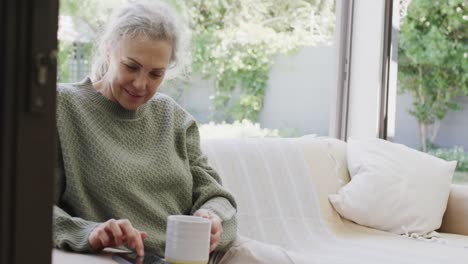 Glückliche-ältere-Kaukasische-Frau,-Die-Auf-Dem-Sofa-Sitzt,-Tee-Trinkt-Und-Ihr-Smartphone-Benutzt,-Zeitlupe