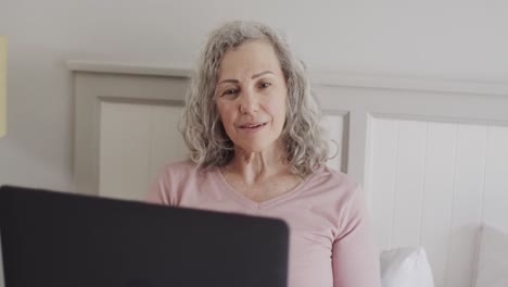 Happy-senior-caucasian-woman-sitting-on-bed-using-laptop,-slow-motion