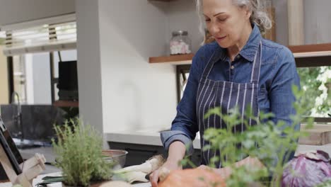 Happy-senior-caucasian-woman-chopping-vegetables-and-using-tablet-in-kitchen,-slow-motion