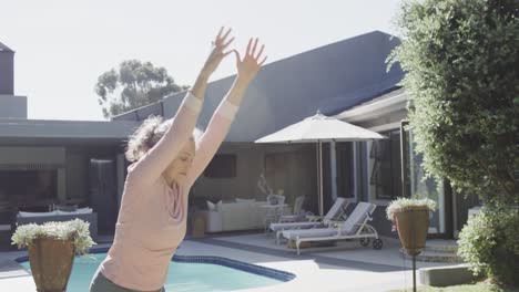 Happy-senior-caucasian-woman-doing-yoga,-stretching-by-swimming-pool-in-garden,-slow-motion