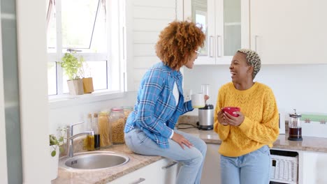 Happy-diverse-female-lesbian-couple-talking-and-drinking-coffee-in-kitchen-in-slow-motion
