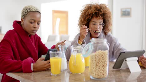 Happy-diverse-female-lesbian-couple-using-tablet-and-having-breakfast-in-kitchen-in-slow-motion