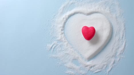 Close-up-of-red-heart-over-sugar-in-heart-shape-on-blue-background-with-copy-space,-slow-motion