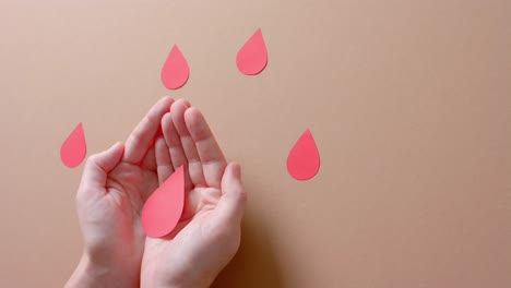 Hands-of-caucasian-woman-holding-blood-drop,-with-drops-on-beige-background,-slow-motion