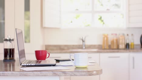 Laptop,-documents-and-mugs-of-coffee-on-countertop-in-kitchen