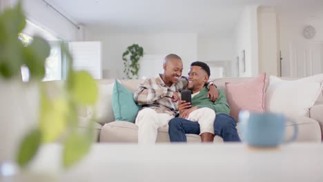Glückliches-Afroamerikanisches-Schwules-Männliches-Paar,-Das-Auf-Dem-Sofa-Sitzt,-Sich-Umarmt-Und-Das-Smartphone-Benutzt,-Zeitlupe
