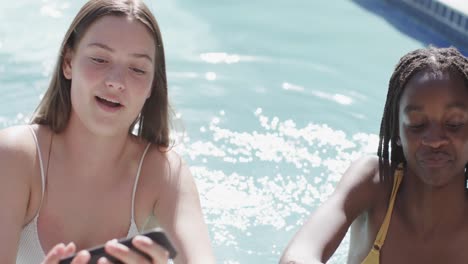 Two-happy-diverse-teenage-female-friends-taking-selfie-in-sunny-swimming-pool-in-slow-motion