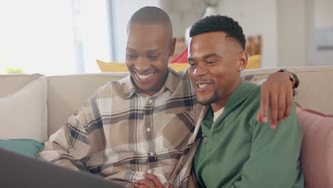 Happy-african-american-gay-male-couple-sitting-on-sofa,-embracing-and-using-laptop,-slow-motion