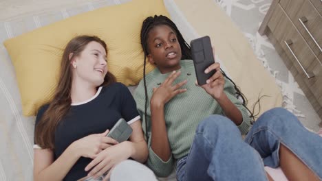 Fröhliche,-Vielfältige-Teenager-Freundinnen,-Die-Mit-Smartphones-Auf-Dem-Bett-Liegen-Und-Lachen