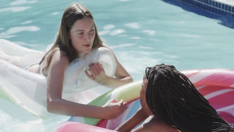 Two-happy-diverse-teenage-female-friends-talking-on-inflatables-in-swimming-pool-in-slow-motion