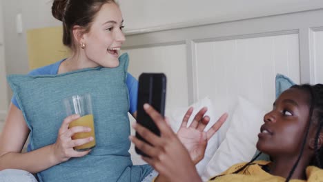 Happy-diverse-teenage-female-friends-lying-on-bed-with-drink,-talking-and-using-smartphone