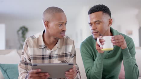 Happy-african-american-gay-male-couple-sitting-on-sofa,-using-tablet,-slow-motion