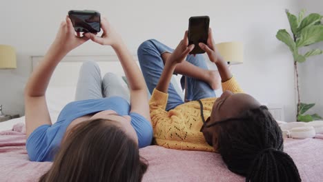 Fröhliche,-Vielfältige-Teenager-Freundinnen,-Die-Auf-Dem-Rücken-Im-Bett-Liegen,-Smartphones-Benutzen-Und-Reden
