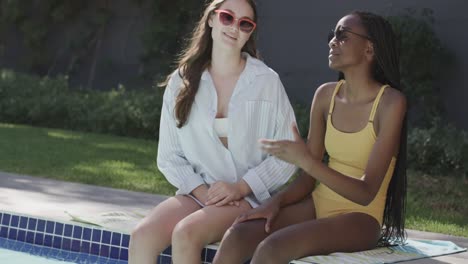 Happy-diverse-teenage-female-friends-in-sunglasses-talking-in-garden-by-pool-in-slow-motion