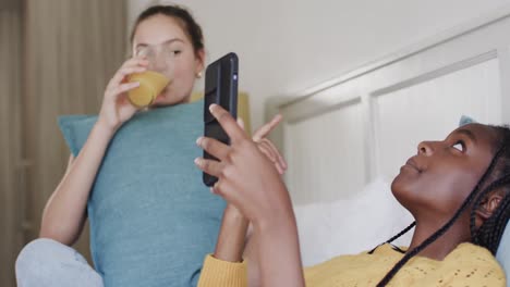 Happy-diverse-teenage-female-friends-lying-on-bed-with-drink,-talking-and-using-smartphone