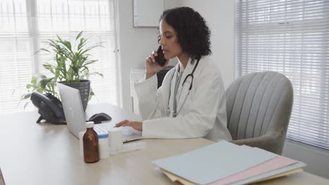 Biracial-female-doctor-sitting-at-desk,-talking-on-smartphone-and-using-laptop,-slow-motion