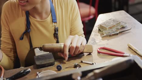 Happy-biracial-female-worker-at-table-shaping-ring-with-handcraft-tools-in-studio-in-slow-motion