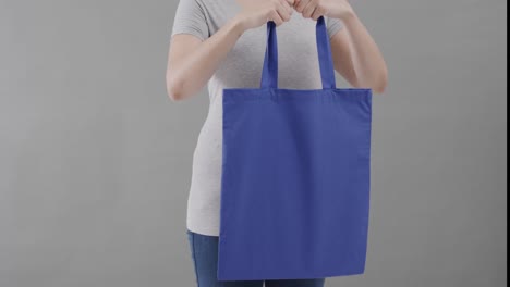 Mujer-Caucásica-Con-Camiseta-Blanca-Sosteniendo-Una-Bolsa-Azul-Sobre-Fondo-Gris,-Espacio-Para-Copiar,-Cámara-Lenta