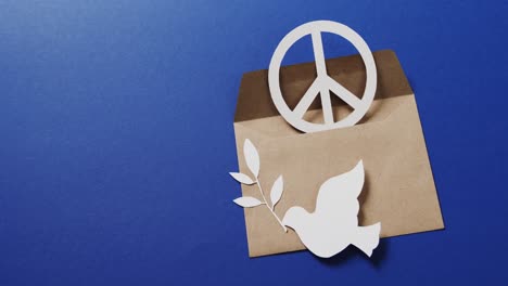 Close-up-of-white-dove-with-leaf,-white-peace-sign,-envelope-and-copy-space-on-blue-background