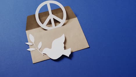 Close-up-of-white-dove-with-leaf,-white-peace-sign,-envelope-and-copy-space-on-blue-background