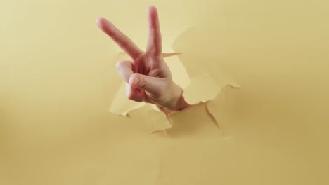 Close-up-of-hand-of-cacausian-woman-showing-peace-sign-with-copy-space-on-torn-yellow-background