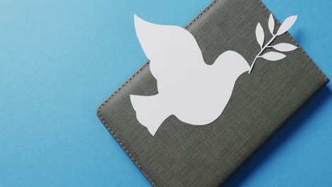 Close-up-of-white-dove-with-leaf-on-grey-notebook-and-copy-space-on-blue-background