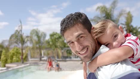 Portrait-of-happy-caucasian-father-carrying-son-at-swimming-pool-at-beach-house