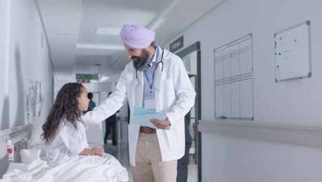 Diverse-sikh-male-doctor-in-turban-and-child-patient-talking-in-corridor-at-hospital,-in-slow-motion