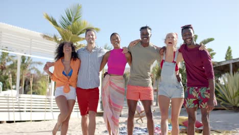 Portrait-of-happy-diverse-group-of-friends-embracing-on-beach-with-beach-house