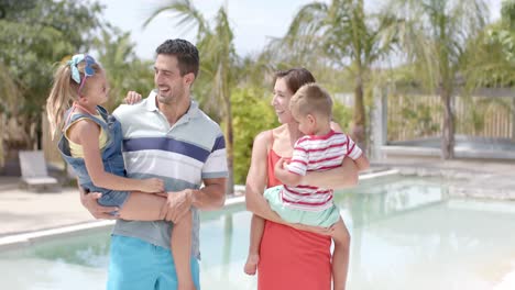 Portrait-of-happy-caucasian-parents-carrying-children-at-swimming-pool-at-beach-house