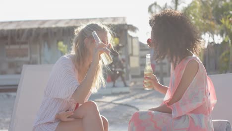 Happy-diverse-female-friends-drinking-beer-and-talking-with-sunglasses-at-beach-house