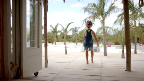 Caucasian-girl-running-and-raising-hands-at-swimming-pool-at-beach-house-with-palm-trees