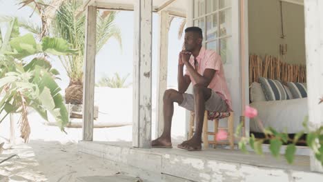 Thoughtful-african-american-man-sitting-on-porch-of-wooden-beach-house-looking-away,-slow-motion