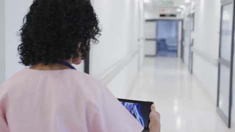 Focused-biracial-female-doctor-walking-and-using-tablet-in-hospital-in-slow-motion