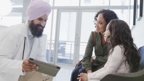 Biracial-sikh-doctor-in-turban-with-tablet-talking-to-girl-with-mother-in-hospital,-slow-motion
