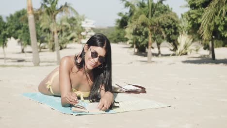 Fröhliche-Hispanische-Frau-Mit-Sonnenbrille,-Die-Am-Strand-Liegt-Und-Ein-Buch-In-Der-Sonne-Liest,-Kopierraum,-Zeitlupe