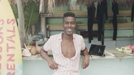 Portrait-of-happy-african-american-man-leaning-and-smiling-at-surfboard-rental-hut,-slow-motion