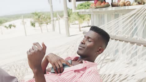 Happy-african-american-man-using-smartphone-lying-in-hammock-on-sunny-beach,-slow-motion