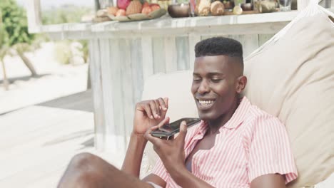 Happy-african-american-man-sitting-in-hammock-talking-on-smartphone-on-sunny-beach,-slow-motion