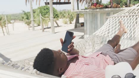 African-american-man-lying-in-hammock-talking-on-smartphone-on-sunny-beach,-slow-motion