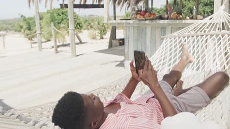 Afroamerikanischer-Mann-Liegt-In-Der-Hängematte-Und-Benutzt-Smartphone-Am-Sonnigen-Strand,-Zeitlupe