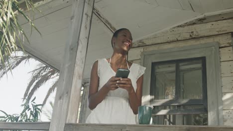 Happy-biracial-woman-in-white-dress-using-smartphone-on-porch-of-wooden-beach-house,-slow-motion
