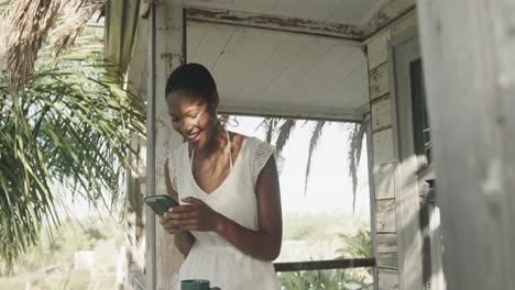 Happy-biracial-woman-in-white-dress-using-smartphone-on-porch-of-wooden-beach-house,-slow-motion