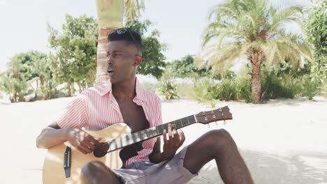 Afroamerikanischer-Mann-Sitzt-Unter-Einem-Baum,-Spielt-Gitarre-Und-Singt-Am-Sonnigen-Strand,-Zeitlupe