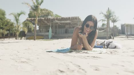 Fröhliche-Hispanische-Frau-Mit-Sonnenbrille,-Die-Am-Strand-Liegt-Und-Auf-Dem-Smartphone-Spricht,-Kopierraum,-Zeitlupe