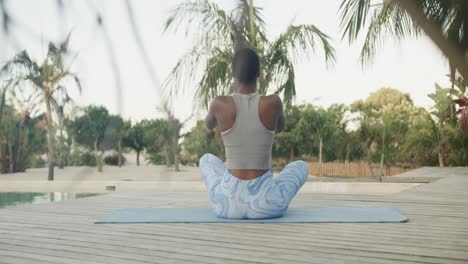 Rückansicht-Einer-Biracial-Frau,-Die-Yoga-Meditation-Praktiziert-Und-Auf-Einem-Steg-Neben-Palmen-Sitzt,-Zeitlupe